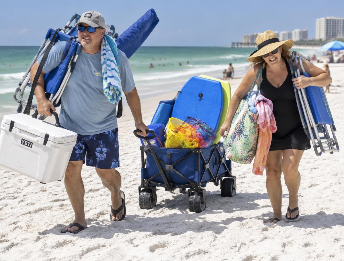 A couple hauling chairs, a cooler, an umbrella and a loaded beach wagon across the white sand of the Florida Gulf Coast